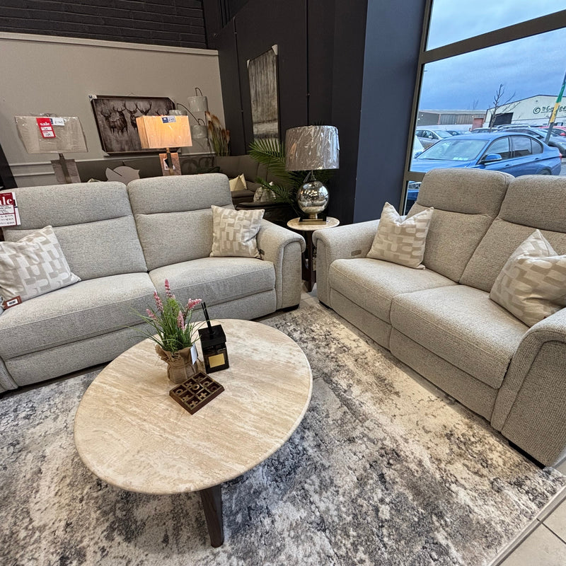 Two Luna Electric Recliners in light gray, each with patterned pillows, are arranged around a round marble coffee table on a textured rug in a modern showroom with large windows and power recline features.