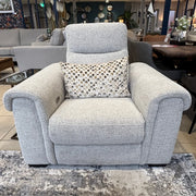 The Luna Electric Recliner, upholstered in light gray fabric and paired with a patterned cushion, is displayed on a gray-patterned rug in a modern showroom alongside contemporary sofas and decorative accents.