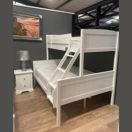 The Karol Bunkbeds, finished in white wood, are displayed in a showroom with gray walls and wood floors. A white nightstand with a silver lamp stands beside them, and a framed landscape photo decorates the wall.