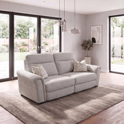 Luna Electric Recliner in light gray fabric, a two-seater sofa with two patterned throw pillows, centered on a rug in a sunlit living room.