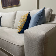 A close-up of the Alice Corner Group in light beige aqua clean fabric, featuring two decorative cushions—one yellow with a floral pattern and one solid blue—set against a plain wall with partially visible framed artwork.