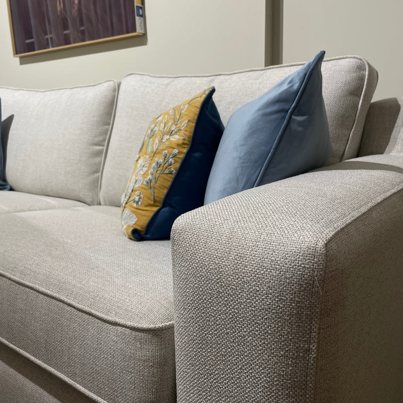 A close-up of the Alice Corner Group in light beige aqua clean fabric, featuring two decorative cushions—one yellow with a floral pattern and one solid blue—set against a plain wall with partially visible framed artwork.