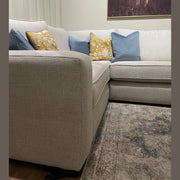 The Alice Corner Group sofa in light gray aqua clean fabric, accented with blue and yellow patterned pillows, sits on a patterned rug. Above the made-to-measure sofa hangs a partial animal painting.