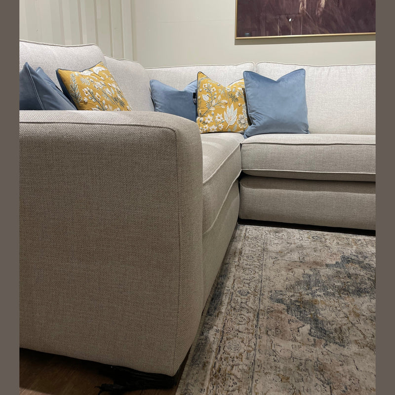 The Alice Corner Group sofa in light gray aqua clean fabric, accented with blue and yellow patterned pillows, sits on a patterned rug. Above the made-to-measure sofa hangs a partial animal painting.