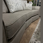 A close-up of the Shelly Angle Seatee, a light gray 4-seater sofa in aquaclean fabric with patterned pillows, shown on a wooden floor near a rug, lamp, and plants in the background.