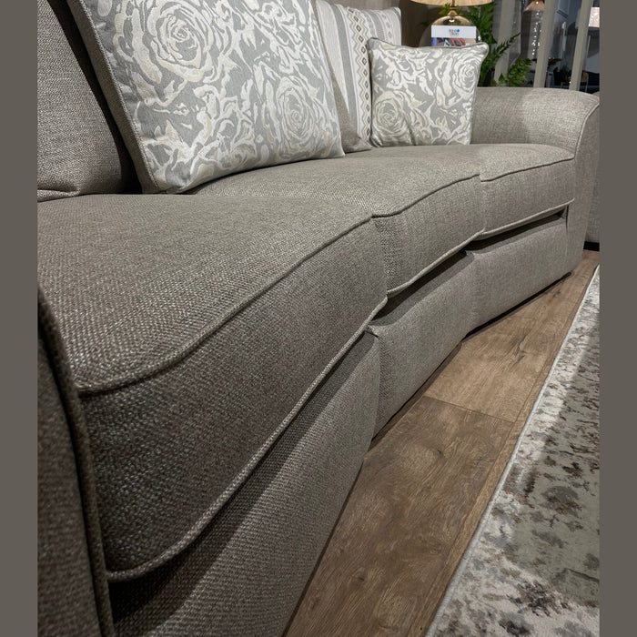 A close-up of the Shelly Angle Seatee, a light gray 4-seater sofa in aquaclean fabric with patterned pillows, shown on a wooden floor near a rug, lamp, and plants in the background.