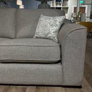 A close-up of the Shelly grey upholstered sofa by vendor-unknown, featuring aquaclean fabric and a floral cushion, displayed in a showroom with wooden flooring and assorted lamps, chairs, and mirrors in the background.