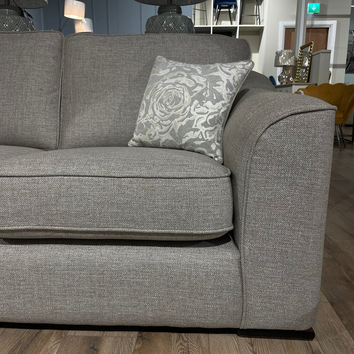 A close-up of the Shelly grey upholstered sofa by vendor-unknown, featuring aquaclean fabric and a floral cushion, displayed in a showroom with wooden flooring and assorted lamps, chairs, and mirrors in the background.