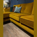 The Turino Chaise, a mustard yellow velvet sectional from our Irish-made corner group, is upholstered in durable aquaclean fabric, shown on a wooden floor in a modern living room with dark blue paneled walls and patterned accent pillows.