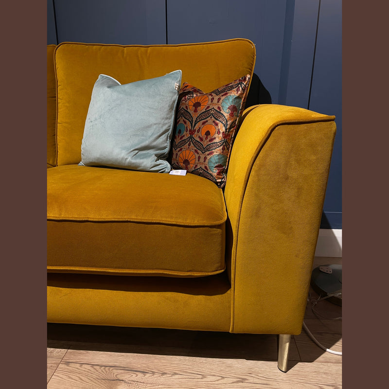 The Turino Chaise, Irish-made with mustard yellow aquaclean velvet, features sleek lines, blue and patterned cushions, and metallic legs, set on a light wooden floor against a dark blue paneled wall.