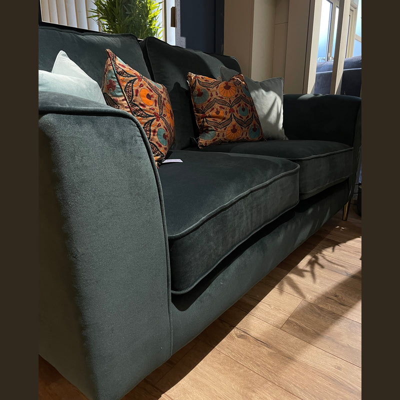 The Turino by vendor-unknown, a dark green velvet corner sofa with patterned cushions, sits on a wooden floor beside a window with sunlight streaming in and a green plant nearby, highlighting its quality and aquaclean fabric charm.