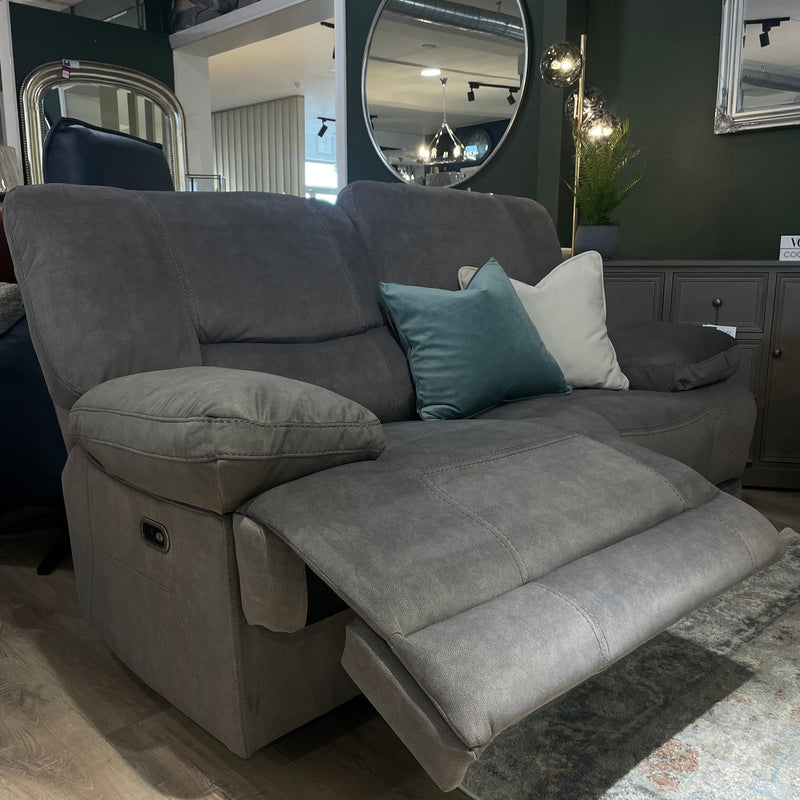 The Gibson Electric Recliner, a modern 3-seater gray sofa with washable fabric, is shown in a showroom with dark green walls, a round mirror, two cushions (teal and white), and a standing lamp in the background.