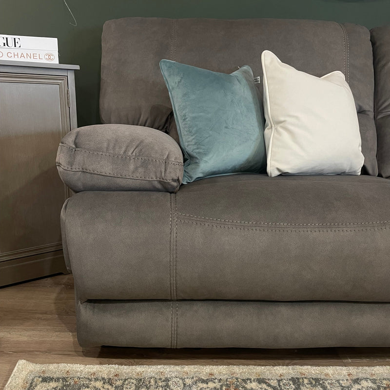 A close-up of the Gibson Electric Recliner in brown suede, styled as a 3-seater with a teal velvet pillow, a cream pillow, and a gray side table. A patterned rug appears on the wooden floor in the foreground.