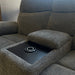 A close-up of the Asher gray electric reclining sofa showcases its central armrest with console storage and a built-in cup holder, as sunlight streams through a nearby window.