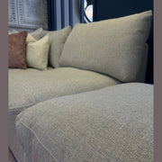 A close-up of the Casablanca Corner Sofa with deep beige seats, three matching cushions, and one brown cushion, set against a dark wall with a decorative panel and part of a rug in the foreground.