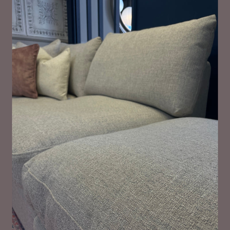 A close-up of the Casablanca Corner Sofa with deep beige seats, three matching cushions, and one brown cushion, set against a dark wall with a decorative panel and part of a rug in the foreground.