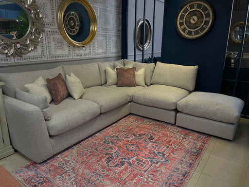 The Casablanca Corner Sofa, light gray and L-shaped with deep seats and beige and brown cushions, rests on a patterned rug. Nearby walls feature decorative mirrors and a large clock, creating a cozy, stylish atmosphere.