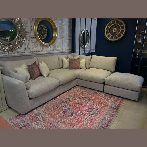 The Casablanca Corner Sofa, light gray and L-shaped with deep seats and beige and brown cushions, rests on a patterned rug. Nearby walls feature decorative mirrors and a large clock, creating a cozy, stylish atmosphere.