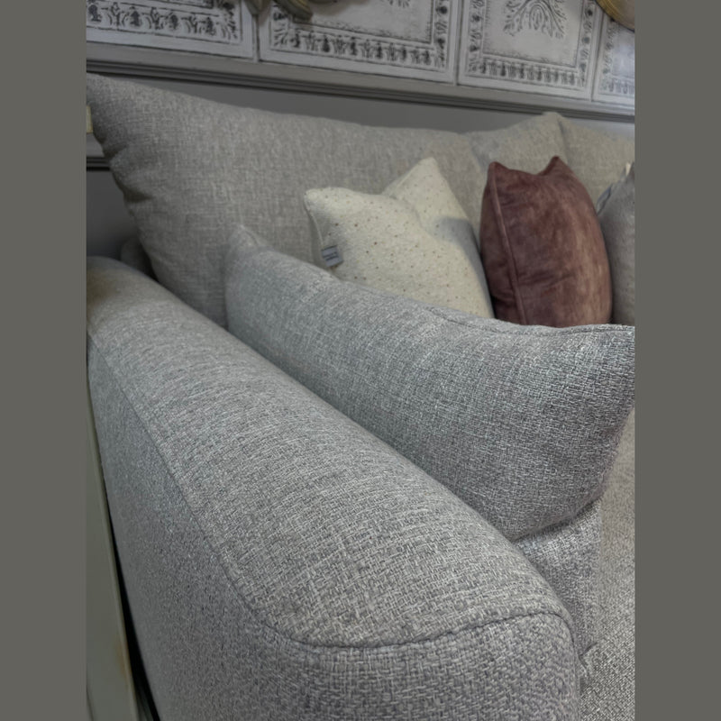 Close-up of the Casablanca Corner Sofa in light gray aqua clean fabric with deep seats, styled with two speckled beige cushions and one mauve velvet pillow, set against a wall featuring decorative white paneling.