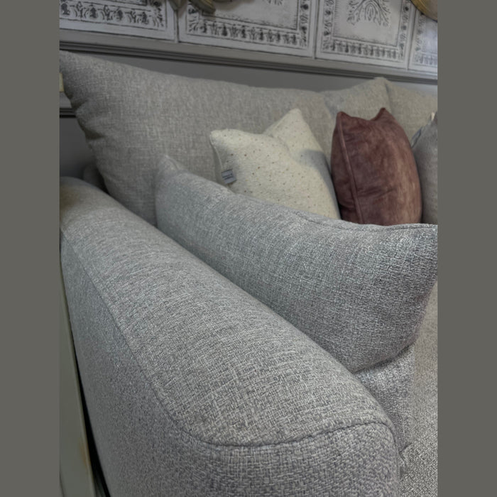 Close-up of the Casablanca Corner Sofa in light gray aqua clean fabric with deep seats, styled with two speckled beige cushions and one mauve velvet pillow, set against a wall featuring decorative white paneling.