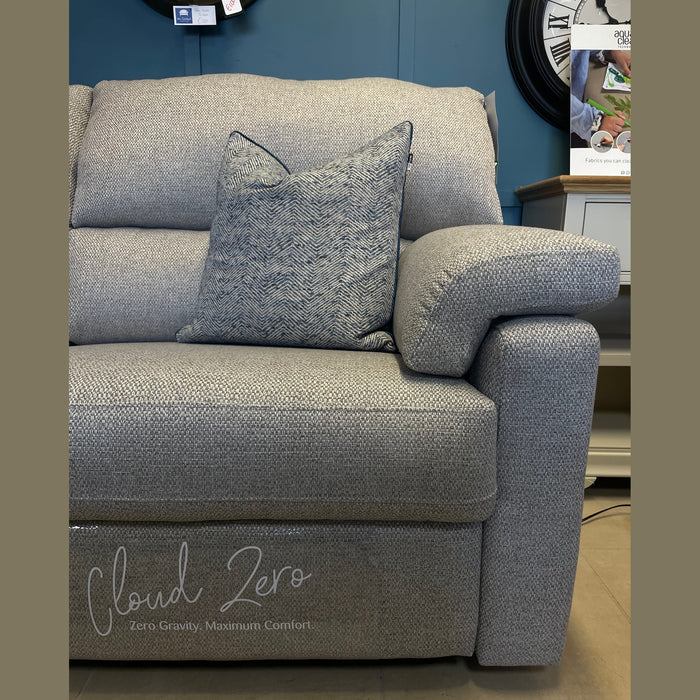 The Hemingway recliner sofa, in light gray aqua clean fabric with a matching cushion, is shown in a showroom. "Cloud Zero, Zero Gravity. Maximum Comfort" is printed on the seat. A white table and clock are in the background.