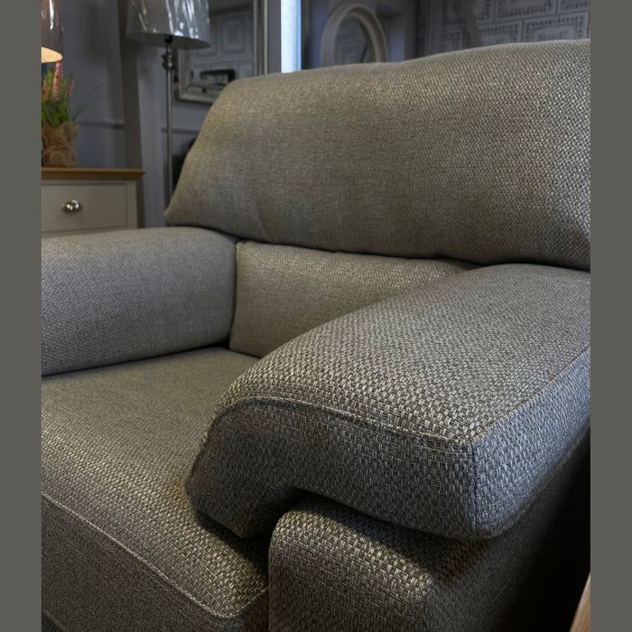 A close-up of the Hemingway gray armchair with premium upholstery in a showroom, featuring a lamp, drawers, and mirrors in the softly lit background.