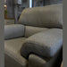 A close-up of the Hemingway gray armchair with premium upholstery in a showroom, featuring a lamp, drawers, and mirrors in the softly lit background.