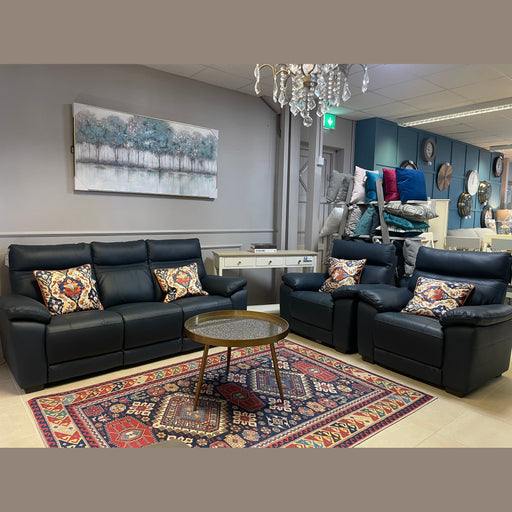 A modern living room display with a black leather Positano Range sofa set by vendor-unknown, colorful patterned pillows, a round glass-top coffee table, a bold geometric rug, wall art with trees, and a reclining armchair in the background.