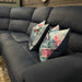 The Dolores Reclining Corner is a dark blue sectional sofa with washable fabric and two floral-patterned pillows in blue, pink, and white, shown against a wooden wall with a mirror and decorative accents in the background.