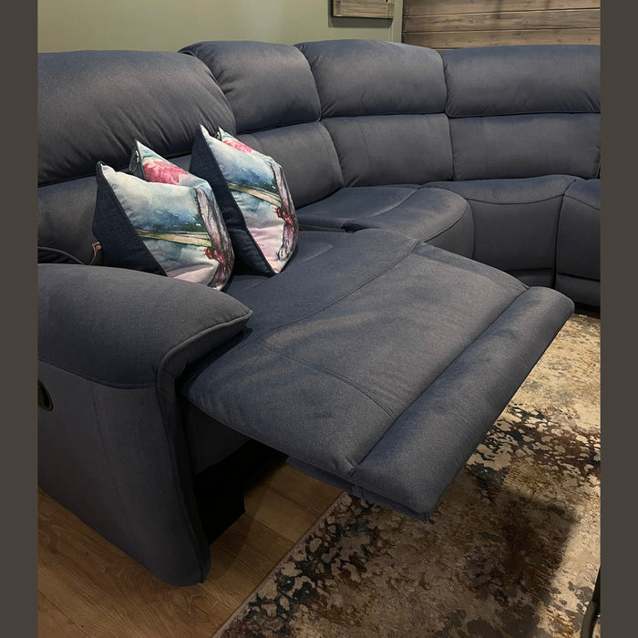 The Dolores Reclining Corner, a modern dark blue recliner sectional sofa, features a reclined footrest, two floral pillows, and sits atop a patterned rug on wooden flooring. A wall with dark wood paneling and a holiday wreath is in the background.