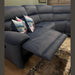 The Dolores Reclining Corner, a modern dark blue recliner sectional sofa, features a reclined footrest, two floral pillows, and sits atop a patterned rug on wooden flooring. A wall with dark wood paneling and a holiday wreath is in the background.