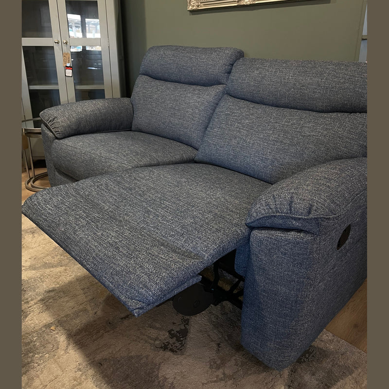 The Vera Recliner Sofa, upholstered in blue with one seat extended, is showcased in a cozy living room featuring a patterned rug, wooden floor, and a cabinet in the background.