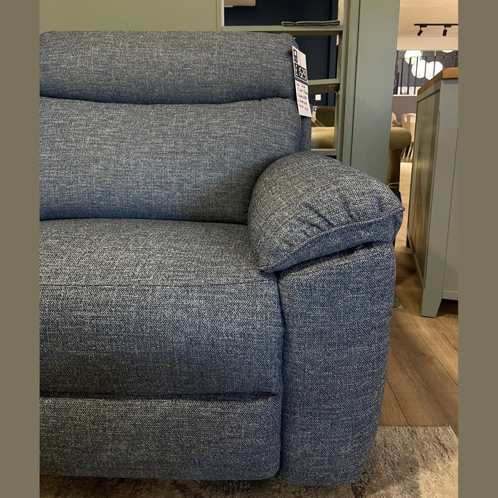 A close-up of the left side of the Vera blue fabric recliner sofa highlights the armrest and backrest, with a small tag on top. The background includes a green wall and wooden shelving, adding to the comfortable suite ambiance.