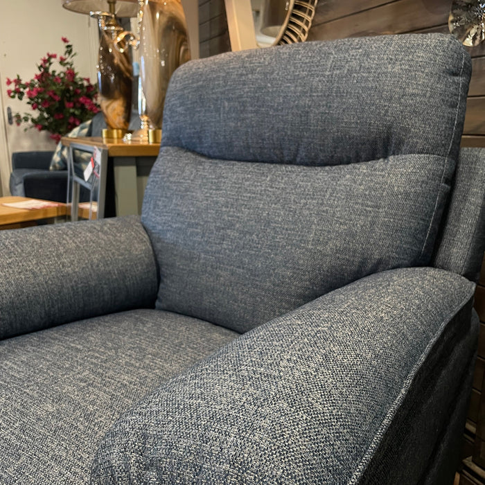 A close-up of the Vera blue upholstered armchair in a cozy, well-lit room with a wooden wall, lamp, round mirror, and flower vase—ideal for bringing comfortable suite vibes and versatile neutral colors to your home décor.