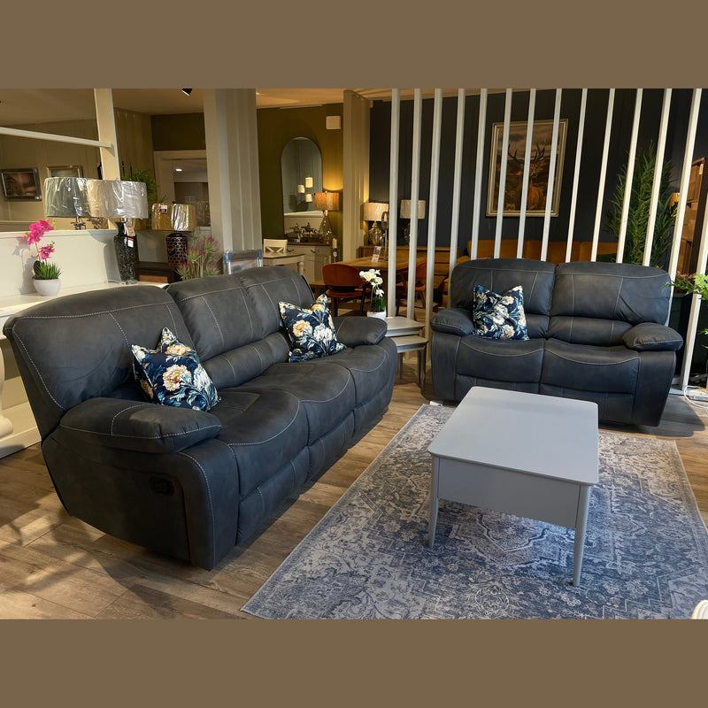 A living room with a Sapphire Reclining Sofa, a dark gray leather sofa, light gray coffee table on a patterned rug, and four floral cushions. The background includes lamps, plants, mirrors, and shelves decorated with décor items.