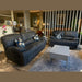 A living room with a Sapphire Reclining Sofa, a dark gray leather sofa, light gray coffee table on a patterned rug, and four floral cushions. The background includes lamps, plants, mirrors, and shelves decorated with décor items.