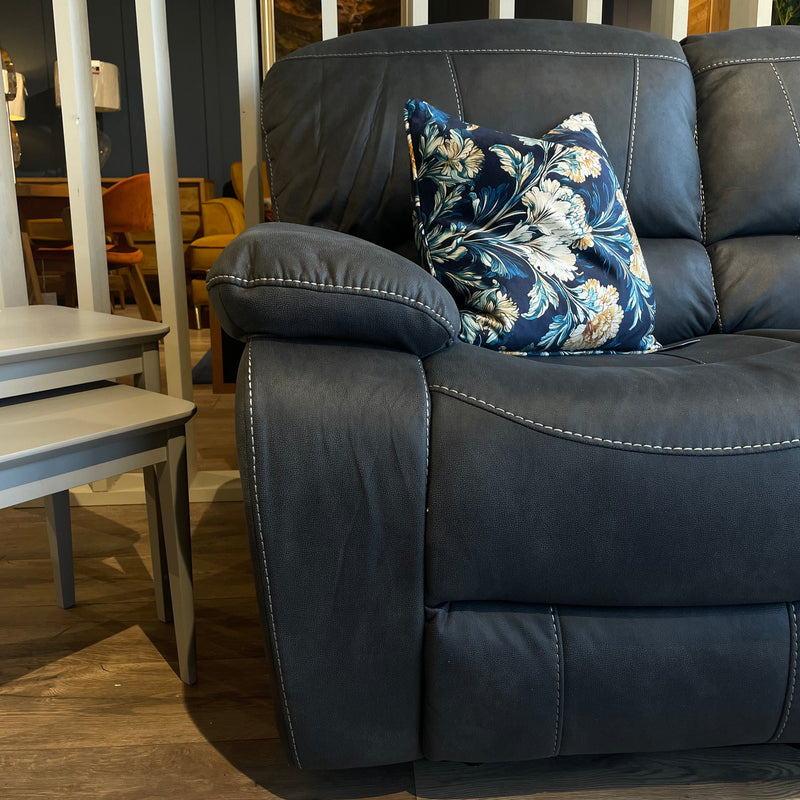 A dark gray Sapphire Reclining Sofa with white stitching, adorned with a blue floral cushion. Two nested light gray side tables are beside the sofa on light wooden flooring.