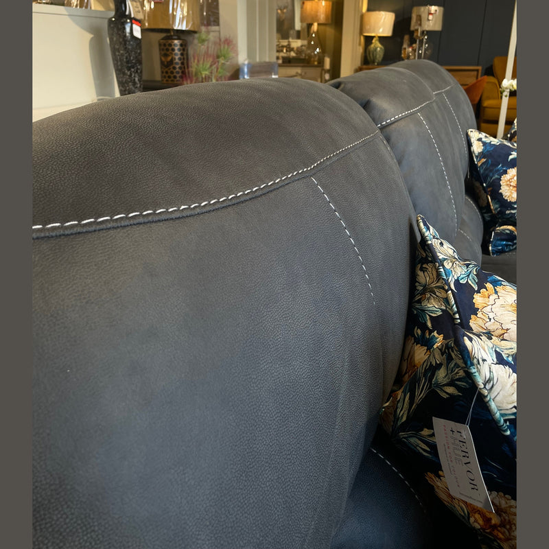 Close-up of the backrest of the Sapphire Reclining Sofa in dark gray durable fabric with white stitching. A floral-patterned cushion is visible, with blurred lamps and decor items in the showroom background.