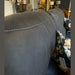 Close-up of the backrest of the Sapphire Reclining Sofa in dark gray durable fabric with white stitching. A floral-patterned cushion is visible, with blurred lamps and decor items in the showroom background.