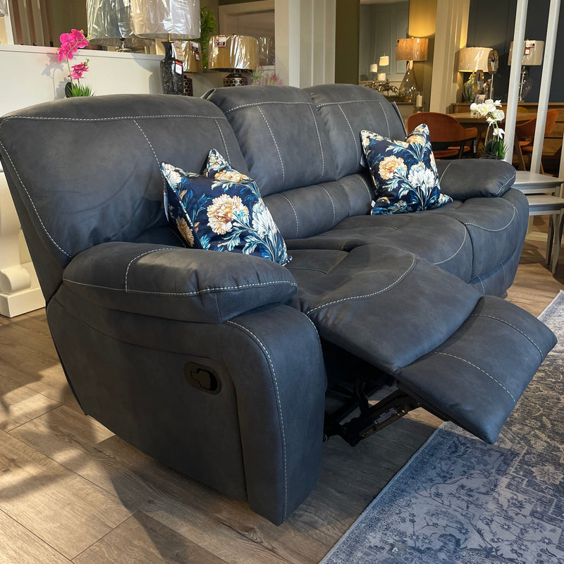The Sapphire Reclining Sofa, upholstered in dark gray durable fabric and paired with floral pillows, is showcased in a showroom on a wooden floor with one footrest extended. Background accents include lamps, flowers, and other furniture.