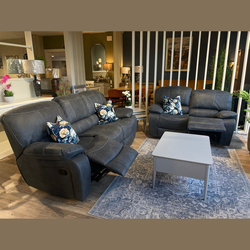 A modern living room featuring two Sapphire Reclining Sofas in dark gray, floral-patterned pillows, a gray coffee table, and a patterned rug on wood flooring. Decorative plants and lamps in the background complete the stylish scene.
