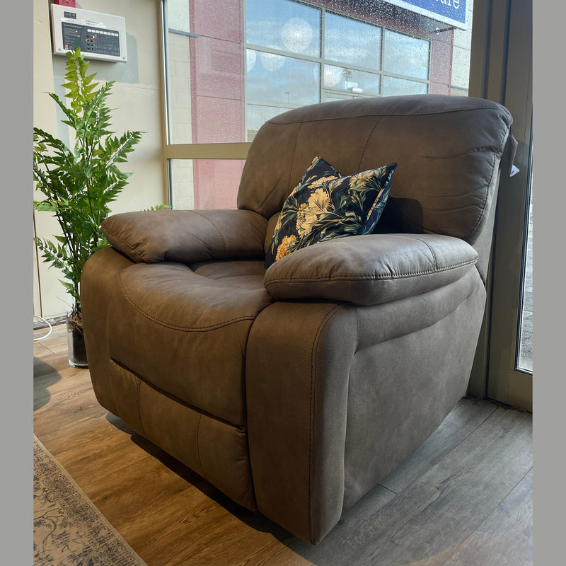 A Sapphire Reclining Sofa is placed by a large window with sunlight streaming in. Two floral decorative pillows rest on the sofa, and a green potted plant sits nearby on the wood floor.