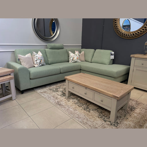 The Harold Chaise Corner, a light green mid century sofa with patterned and beige pillows, sits in a modern living room with a light wood coffee table featuring drawers. Decorative mirrors hang on gray and white walls behind.