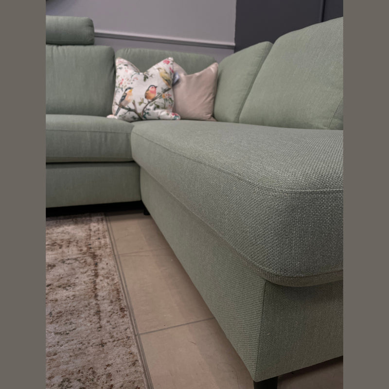 A close-up of the Harold Chaise Corner sectional sofa in light green, styled with a floral and a pale pink pillow, placed beside a beige carpet and tiled floor against gray walls.