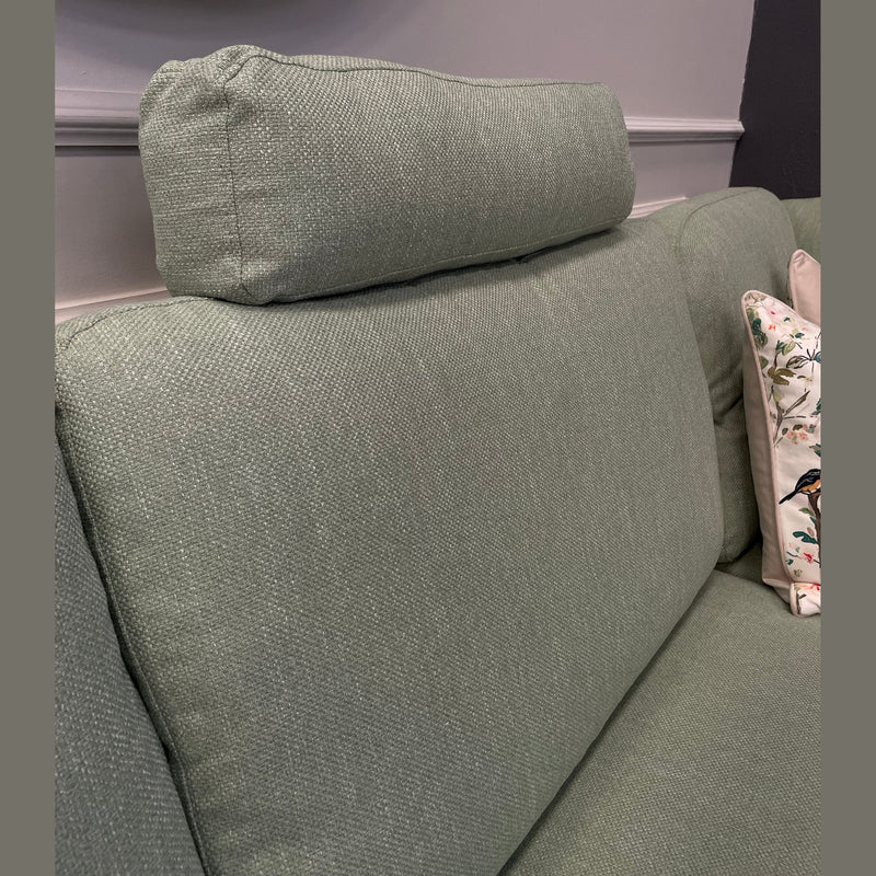 Close-up of the Harold Chaise Corner, a light green mid-century fabric sofa with a cylindrical headrest and a decorative cushion featuring birds and florals. A round mirror and grey wall are seen in the background.