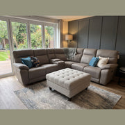A gray leather Jude Corner Sofa with a center console and matching tufted ottoman on a patterned area rug.