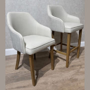 A standard modern chair with cushioned seat and wooden legs sits beside a Como Counter Stool in neutral fabric, which stands taller on the wooden floor; both are arranged against a light-colored wall.