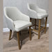 A standard modern chair with cushioned seat and wooden legs sits beside a Como Counter Stool in neutral fabric, which stands taller on the wooden floor; both are arranged against a light-colored wall.