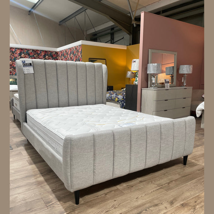 The Isobella Bedframe, upholstered in light gray stone linen with a high vertically tufted headboard, is showcased in a showroom with wooden floors, colorful accent walls, and a dresser topped with matching lamps.