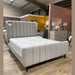 The Isobella Bedframe, upholstered in light gray stone linen with a high vertically tufted headboard, is showcased in a showroom with wooden floors, colorful accent walls, and a dresser topped with matching lamps.